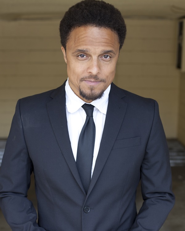 A picture of voice actor Catero Colbert, a young Black man with short facial hair, wearing a suit and tie.