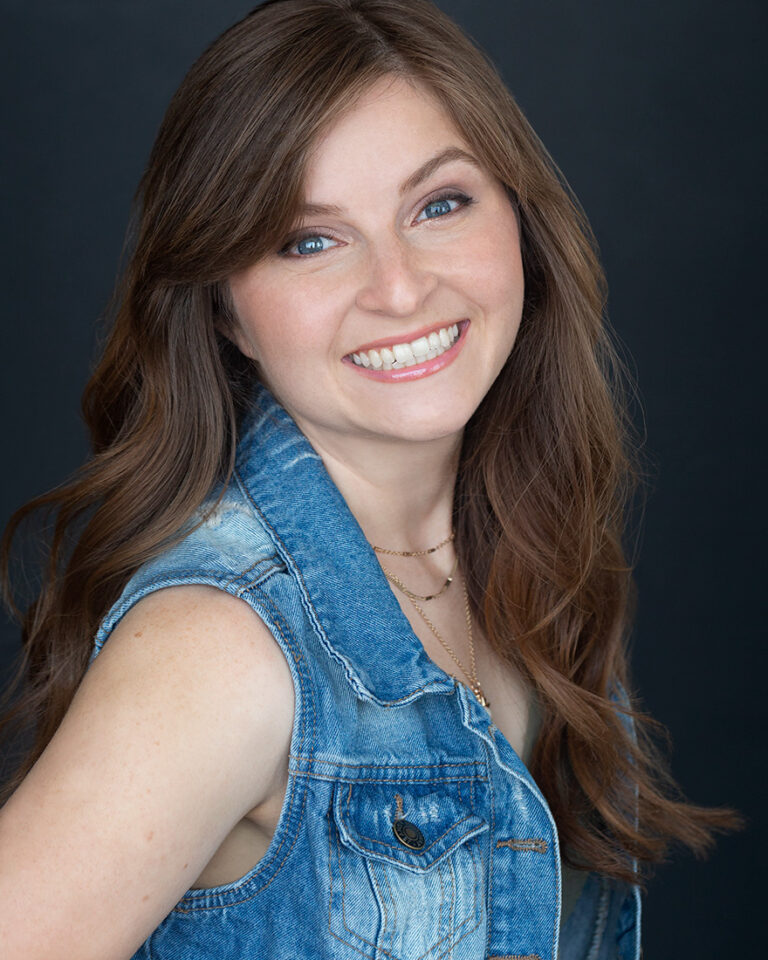 A picture of voice actor Lizzie Freeman, a young white woman with medium length brown hair, wearing a denim vest.